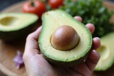 Primo piano di una mano che tiene un avocado tagliato a metà, mettendo in evidenza la sua polpa verde e il nocciolo, con uno sfondo sfocato di altri ingredienti freschi.